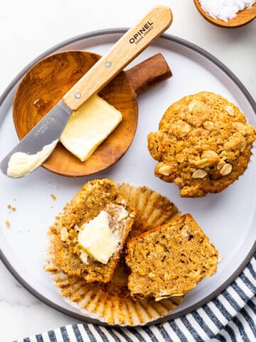 A grey-rimmed ceramic plate with two banana muffins, one split open and buttered.