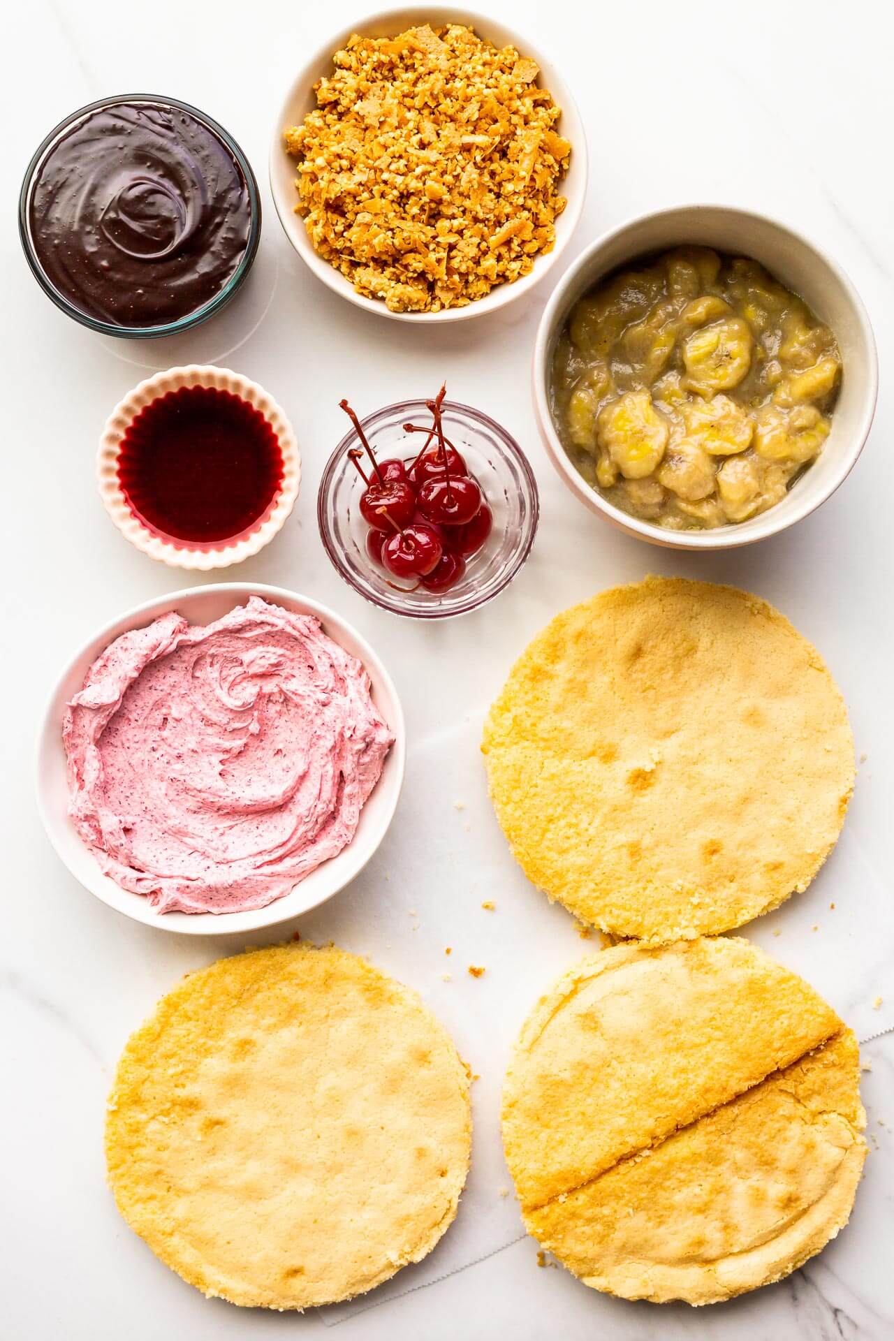 The components of a banana split cake, ready to be stacked and assembled into a layer cake, including three layers of vanilla cake, cherry frosting, maraschino cherries, cherry juice, banana compote, peanut crunch, and chocolate fudge sauce.