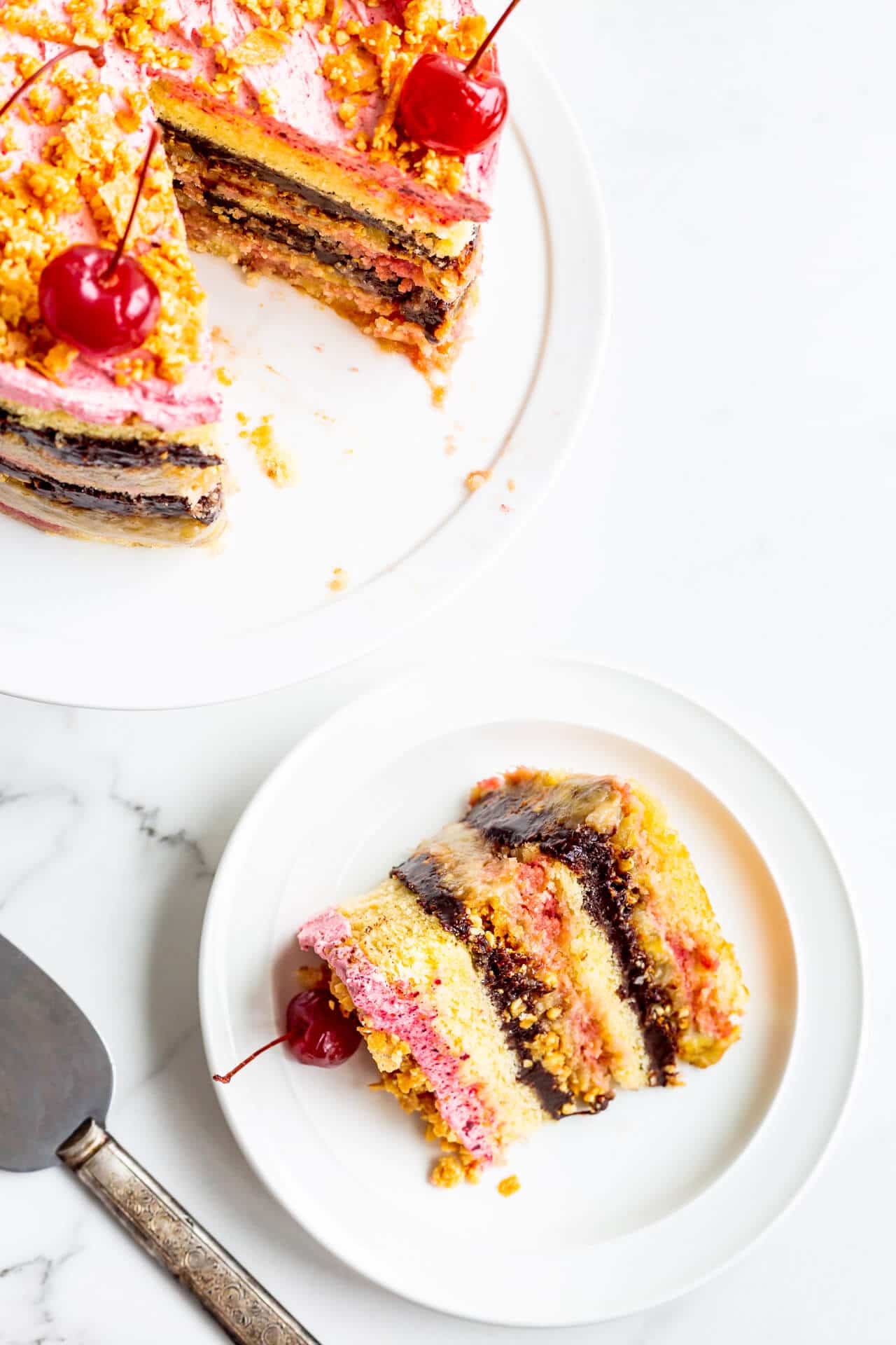 Banana split layer cake topped with maraschino cherries on a cake stand being served on plates.