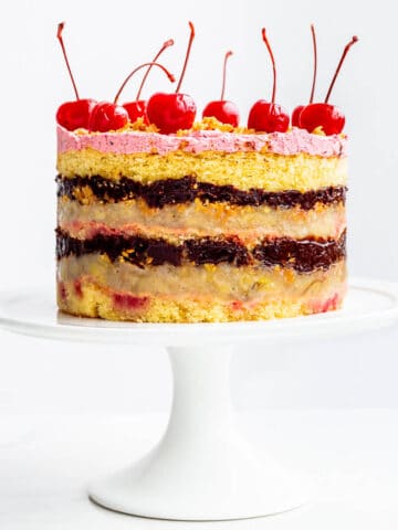 Banana split layer cake with layers of chocolate fudge filling and banana, and maraschino cherries on a white cake stand.