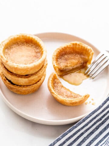 Butter tarts stacked on a beige plate with one cut open with a fork to reveal the gooey filling.