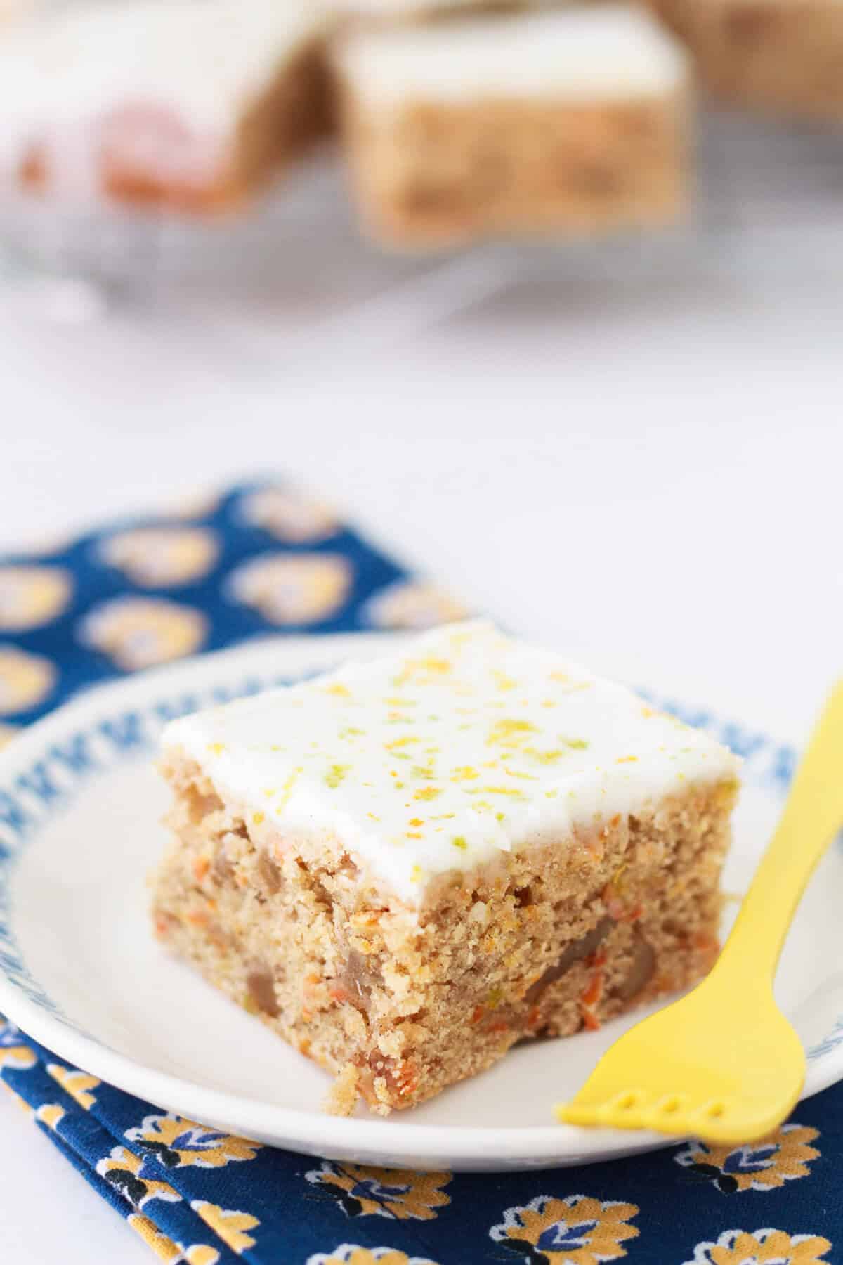 A square of carrot cake on a dessert plate with a yellow fork.