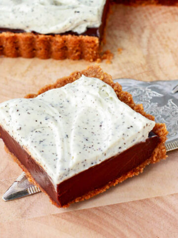 A square piece of a chocolate tart topped with whipped cream on a wooden board.