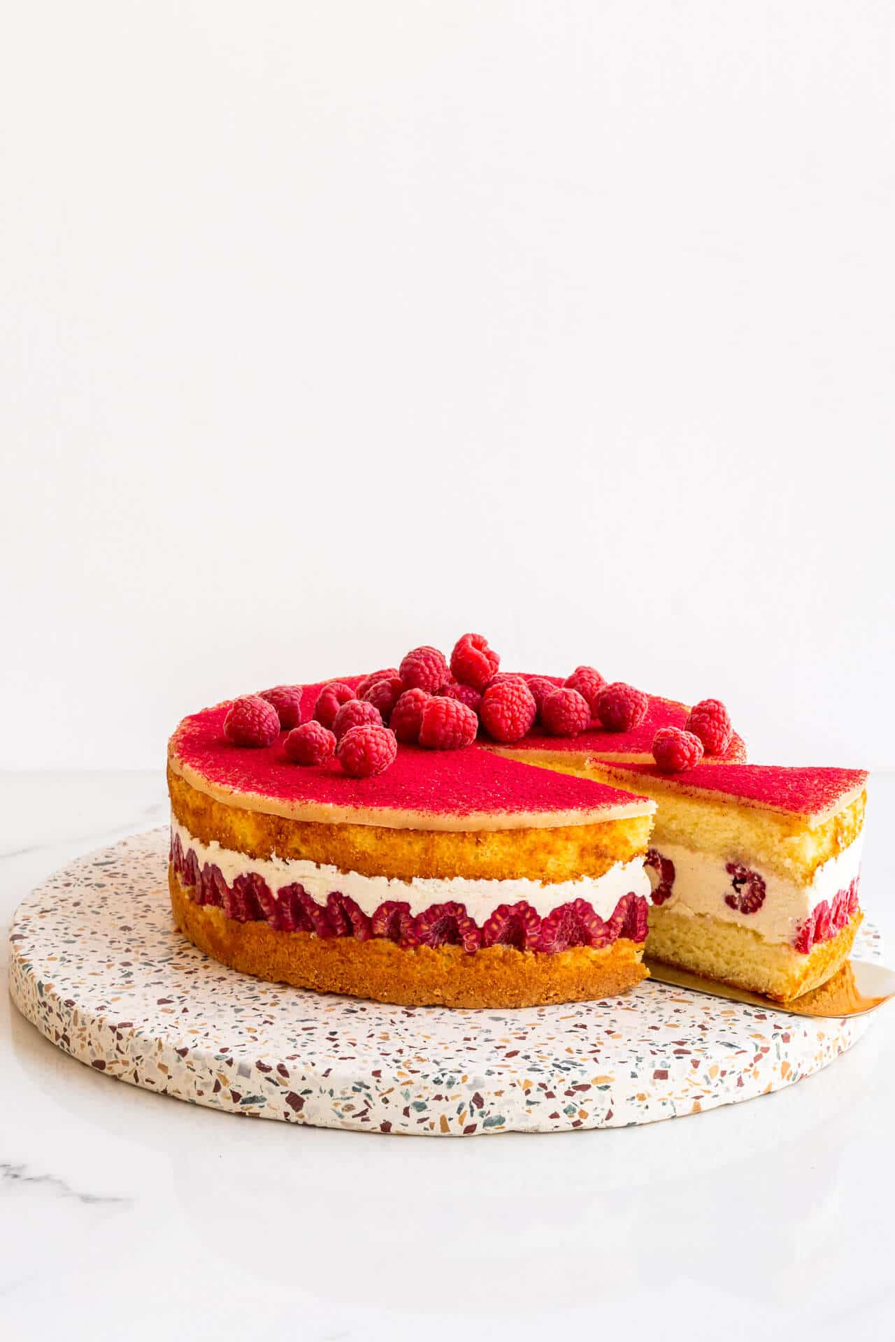 Framboisier cake topped with fresh raspberries on a terrazzo board being served with a gold cake lifter.