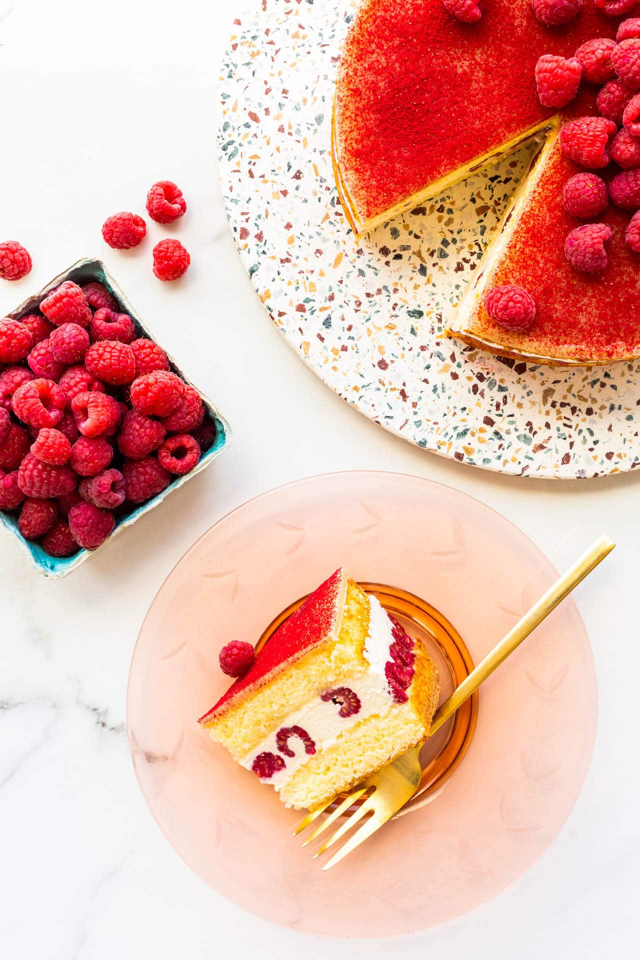 A slice of framboisier cake on a pink glass plate with a gold fork.