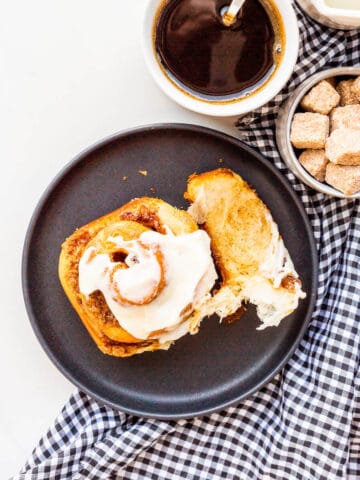 A cinnamon raisin bun on a black plate being eaten, with a cup of coffee on the side.