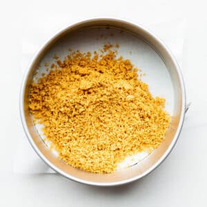 Graham cracker crumbs in a springform pan lined with parchment paper, to be spread out and pressed to form a crust for cheesecakes.
