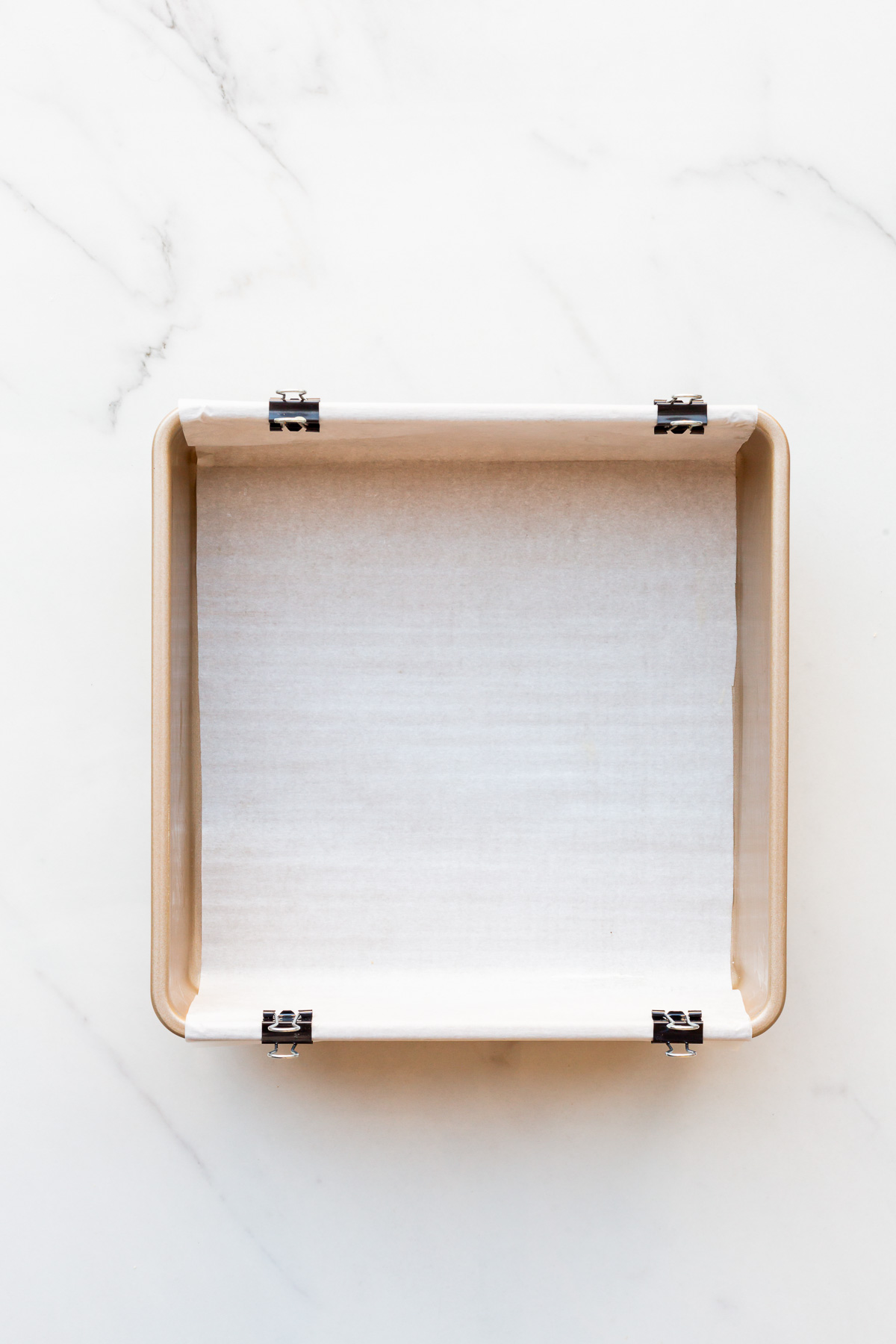 A square metal baking pan lined with parchment paper with an overhang secured in place with binder clips.