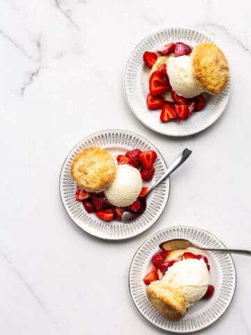 Three ice cream strawberry shortcakes on small dessert plates on white marble surface