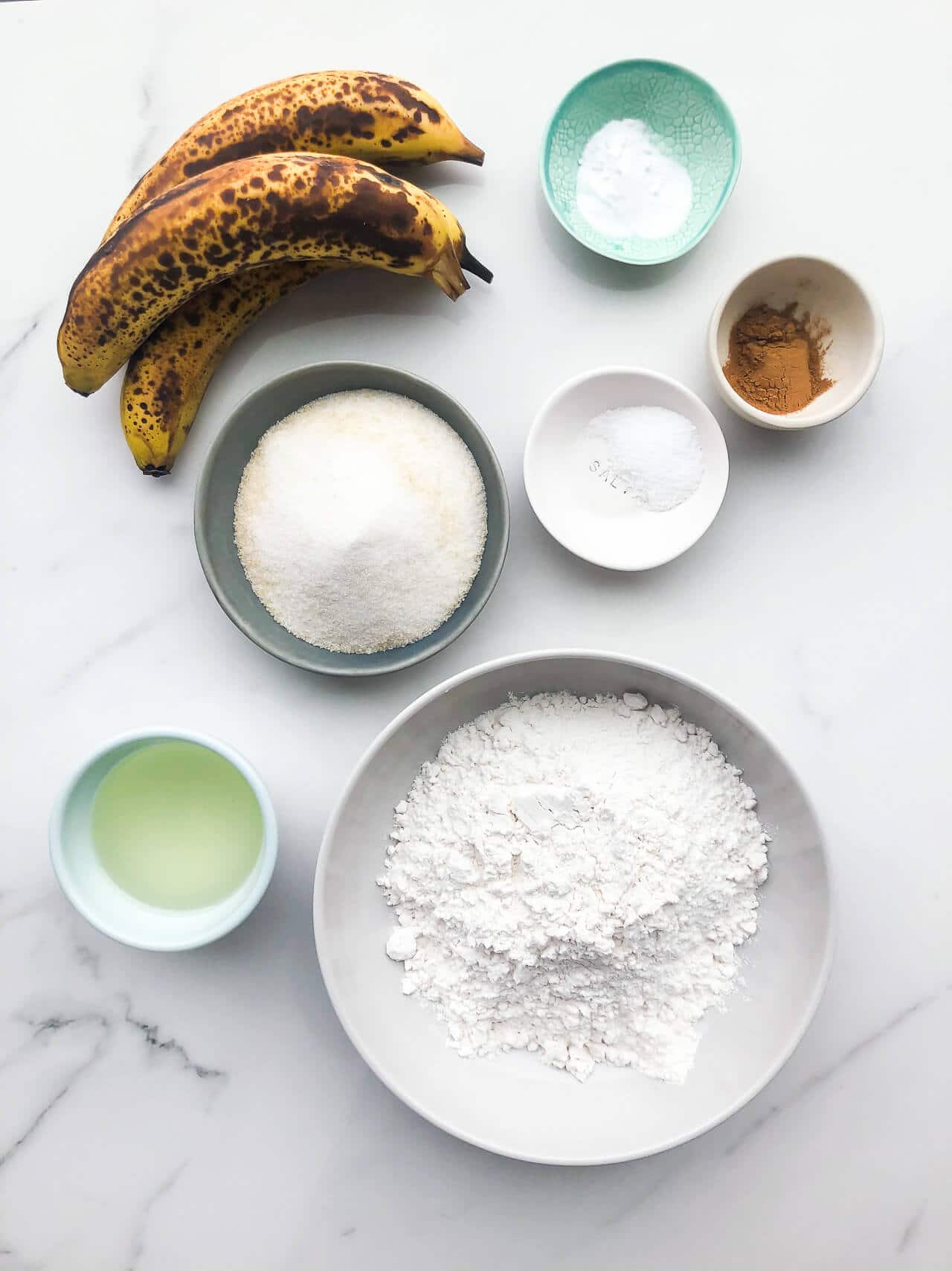 Bowls of ingredients for vegan banana bread (eggless and dairy-free) include oil, bananas, sugar, flour, baking soda, salt, cinnamon