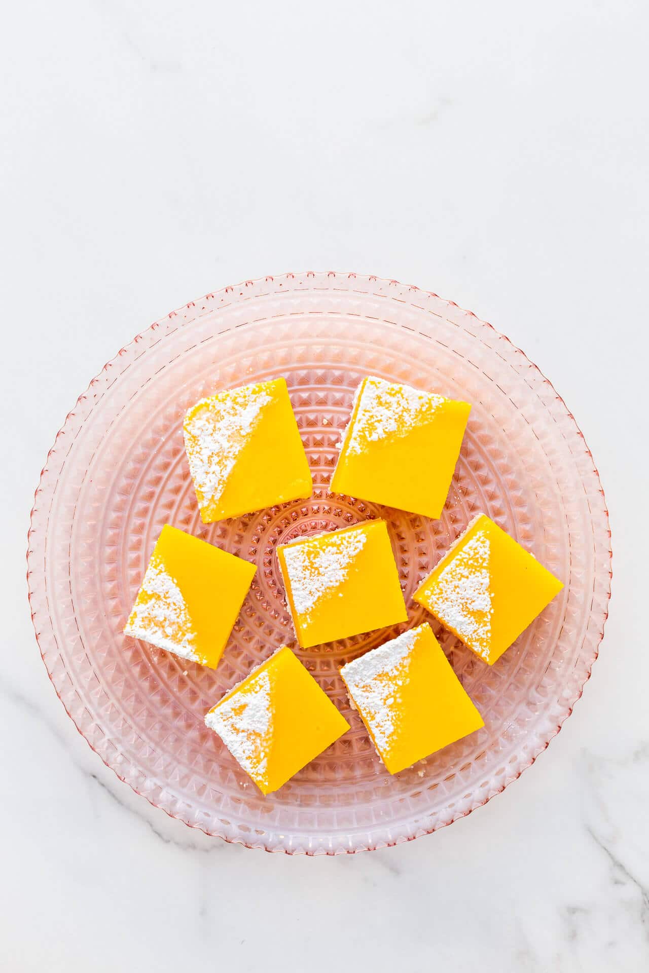 A pink glass plate with lemon bars topped with powdered sugar, ready to serve.