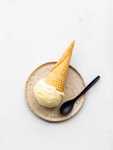 A scoop of lemon ice cream served on a speckled plate with a cone and a black spoon