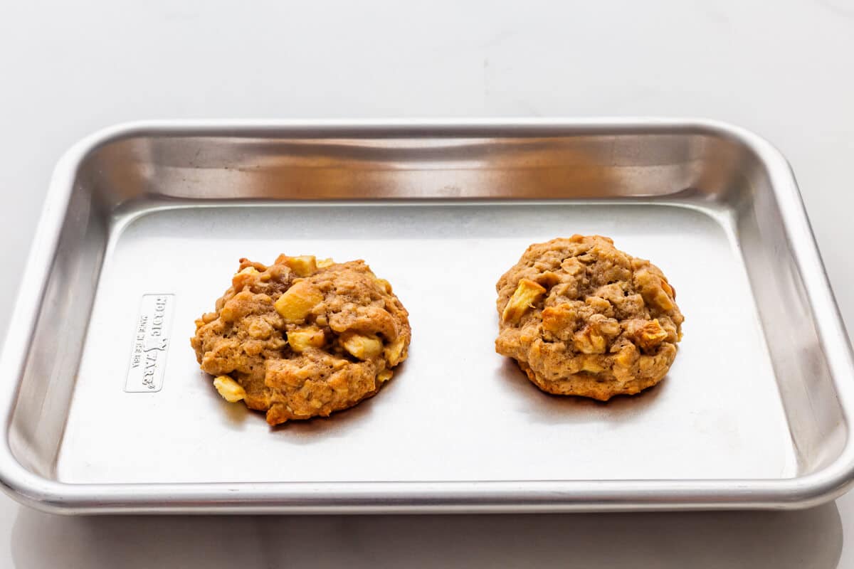 Two oatmeal cookies on a mini sheet pan. One has spread out more than the other as it baked so is wider.