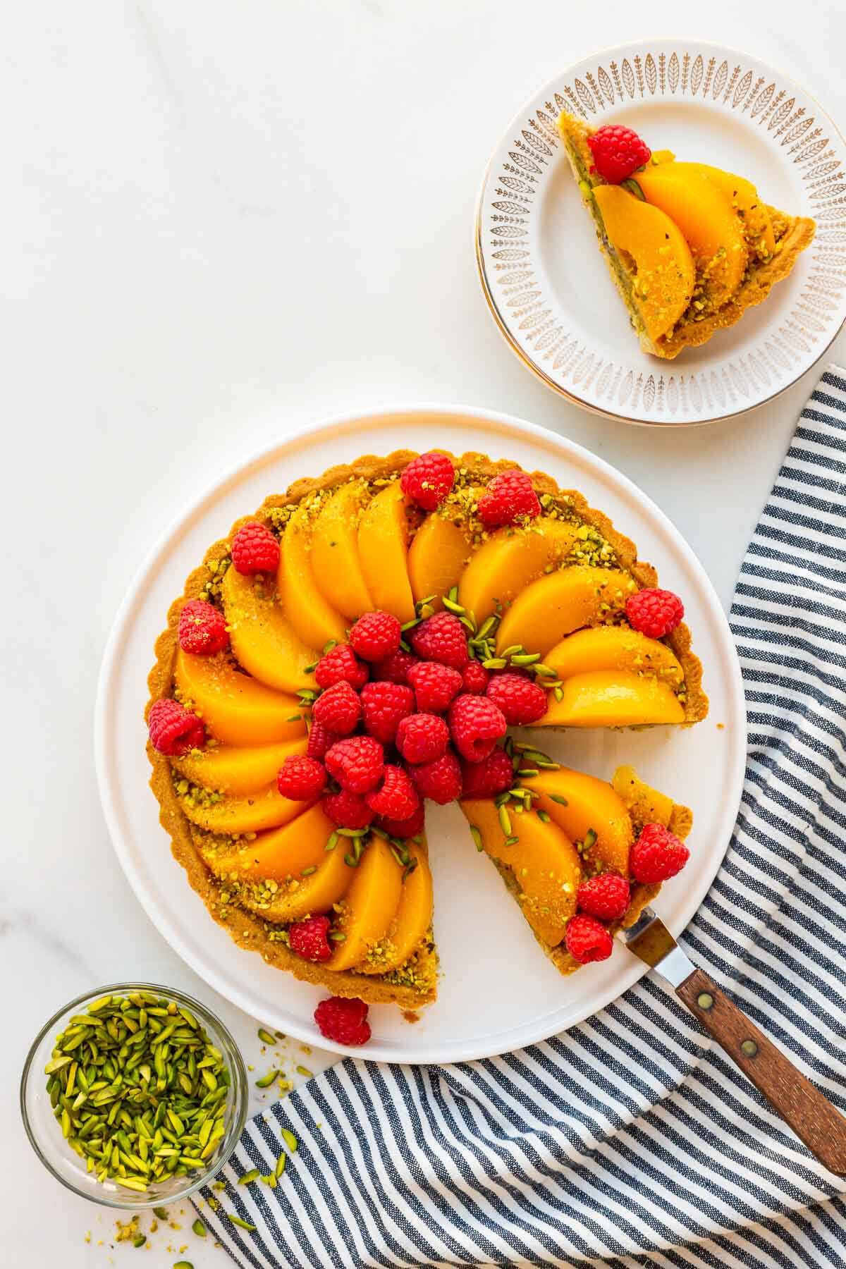 Slicing and serving a pistachio tart topped with sliced peaches and fresh raspberries, and decorated with slivered and ground green pistachios.