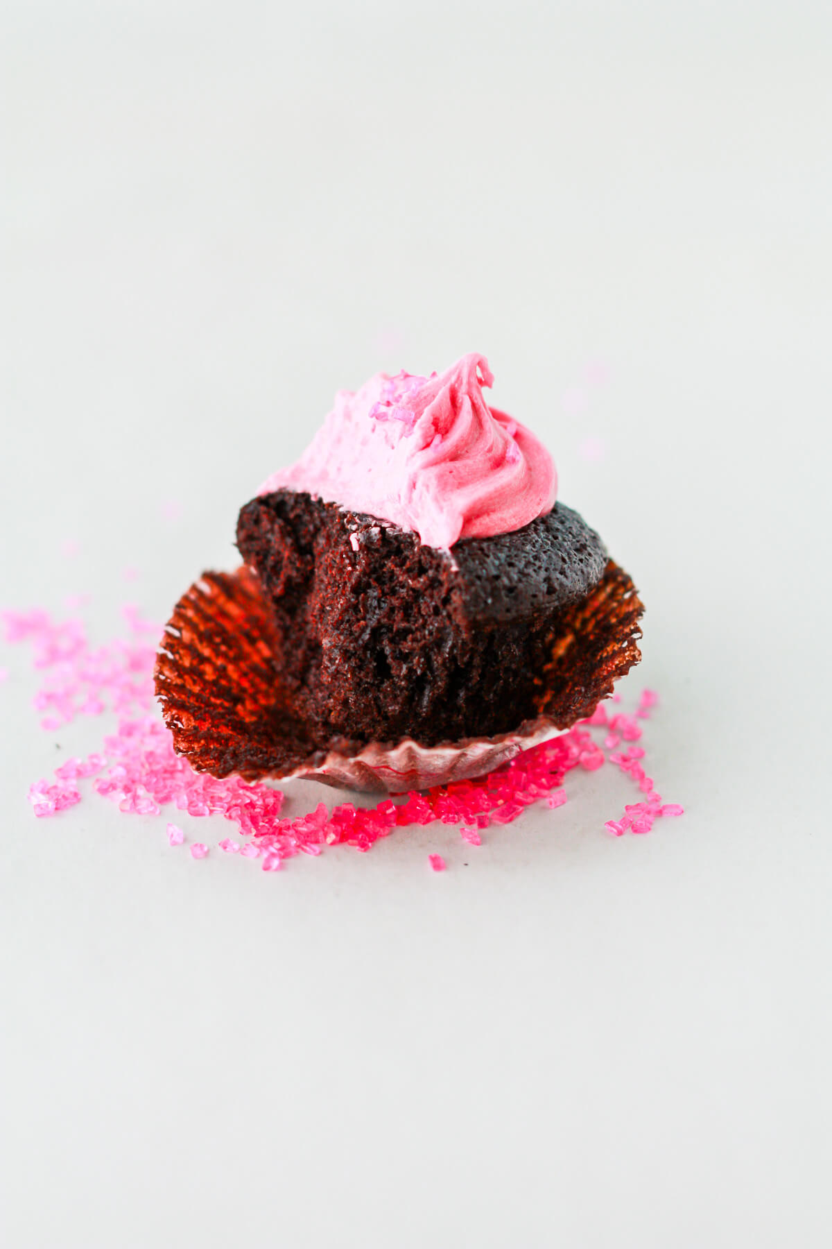 A dark chocolate cupcake with pink frosting bitten into to show the moist crumb.