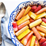 A blue and white round ceramic baking dish with roasted pink rhubarb and a few strawberries in pink syrup with blue and white striped linen and big vintage serving spoon