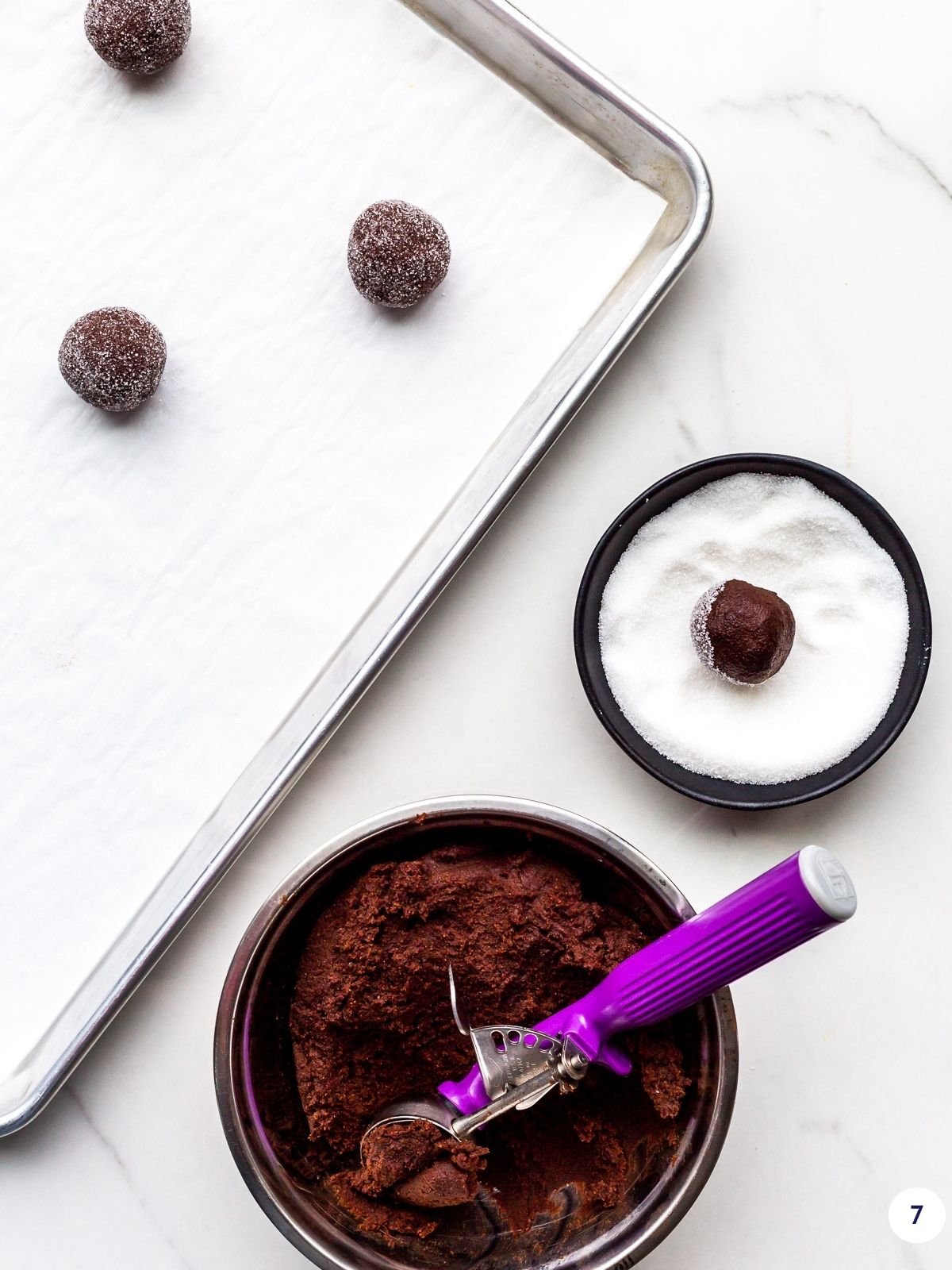 Scooping chocolate sugar cookie dough and then rolling in granulated sugar before baking on a sheet pan.