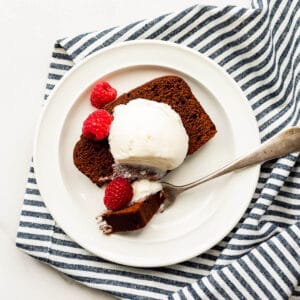 Slice of chocolate loaf cake with a scoop of ice cream and fresh raspberries on a plate with a fork, with a striped linen tucked underneath.