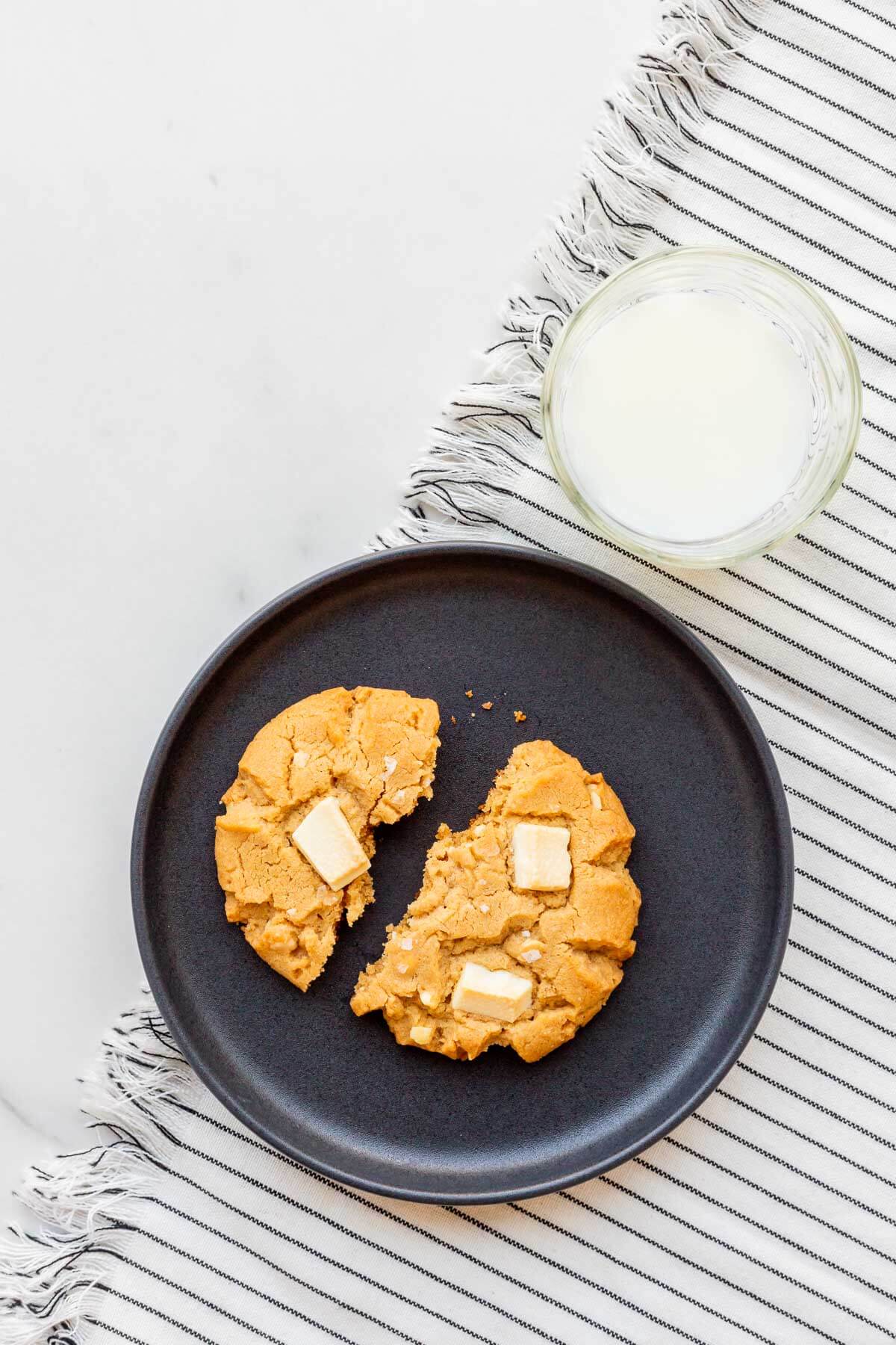 A big peanut butter cookie on a plate broken in half with a glass of milk on the side.