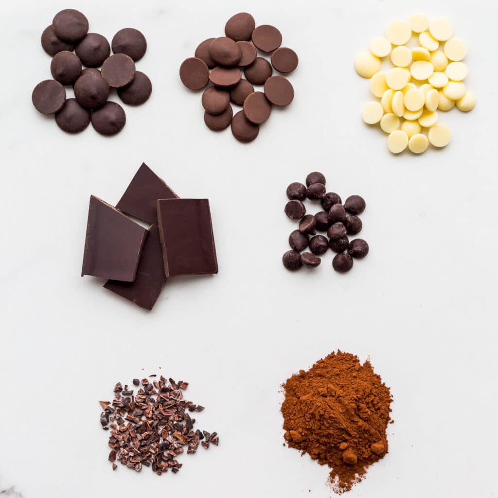 Different types of chocolate (chocolate pistoles, chocolate chips, milk, white, and dark chocolate, cocoa nibs and cocoa powder) set on a white marble surface.