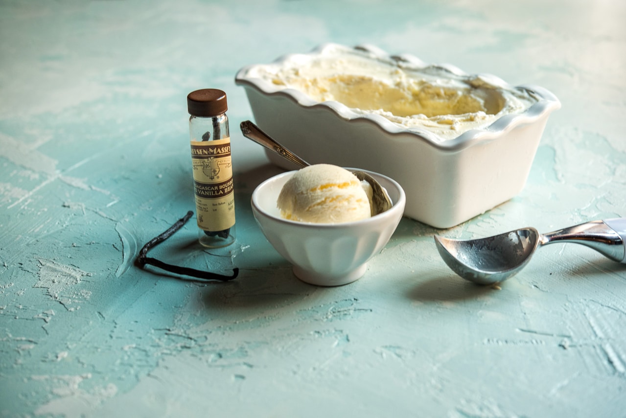 White tub of vanilla bean ice cream served in white bowl next to jar of vanilla beans and ice cream scoop