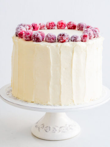 A white chocolate cranberry cake topped with sugared cranberries on a cake stand.